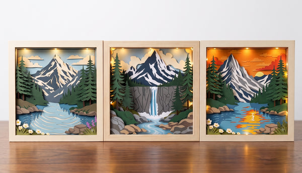 Three framed layered wood shadow box art prints depicting mountain landscapes with snowy peaks, pine trees, rivers, and waterfalls, arranged side-by-side on a wooden surface against a white wall.